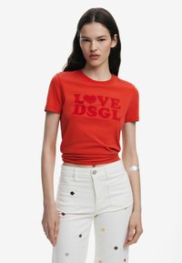 Rotes Baumwoll-T-Shirt mit kurzen Ärmeln, versehen mit "LOVE" und "DSGL" in strukturierter Stickerei. Das Modell kombiniert es mit weißen Jeans, die mit bunten Blumenstickereien verziert sind.