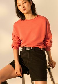 Koraliowa, oversize'owa bluza z podwiniętymi mankietami, zestawiona z czarnymi szortami z wysokim stanem i srebrną klamrą, na jasnym tle.