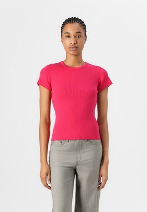 Kurzärmliges, tailliertes T-Shirt aus leuchtend pinkem Rippstoff. Rundhalsausschnitt, glatte Textur, kombiniert mit hellgrauen Hosen.