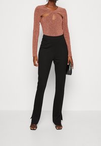 Kvinna klädd i en rostfärgad långärmad topp med urtag, svarta högmidjade byxor, svarta sandaler och håller en svart kuvertväska.