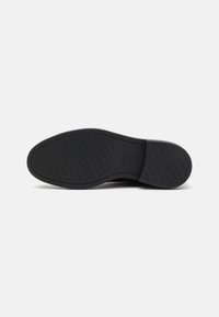 Black rubber shoe sole with textured grip pattern, curved edges, and embossed branding. Flat and slightly tapered shape.