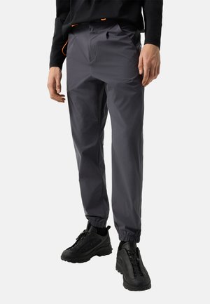 Mand iført mørkegrå joggingbukser med elastiske manchetter og sorte sportssko, stående mod en ensartet lys baggrund.