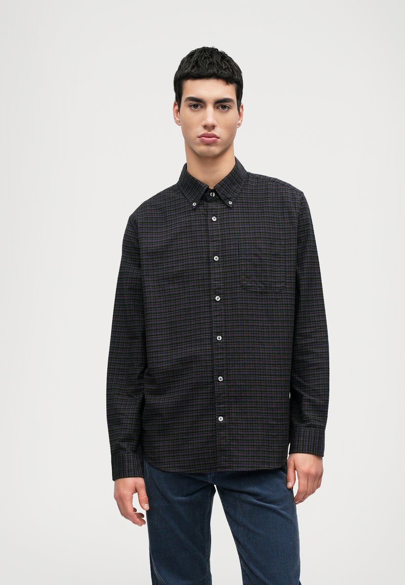 Fekete és lila kockás, hosszú ujjú ing gombolható gallérral, elülső zsebbel és ezüst gombokkal; sötétkék textúrált nadrággal viselve.