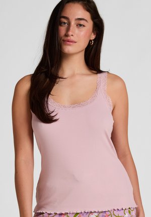 Jonge vrouw met lang donker haar die een roze top met kanten rand en een broek met bloemenpatroon draagt, poserend tegen een effen achtergrond.