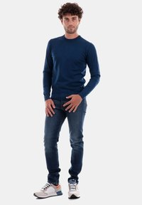 Camicia a maniche lunghe blu navy, jeans blu e scarpe leggere con accenti crema, grigio e arancione. Il modello si presenta con le mani posizionate sui fianchi.