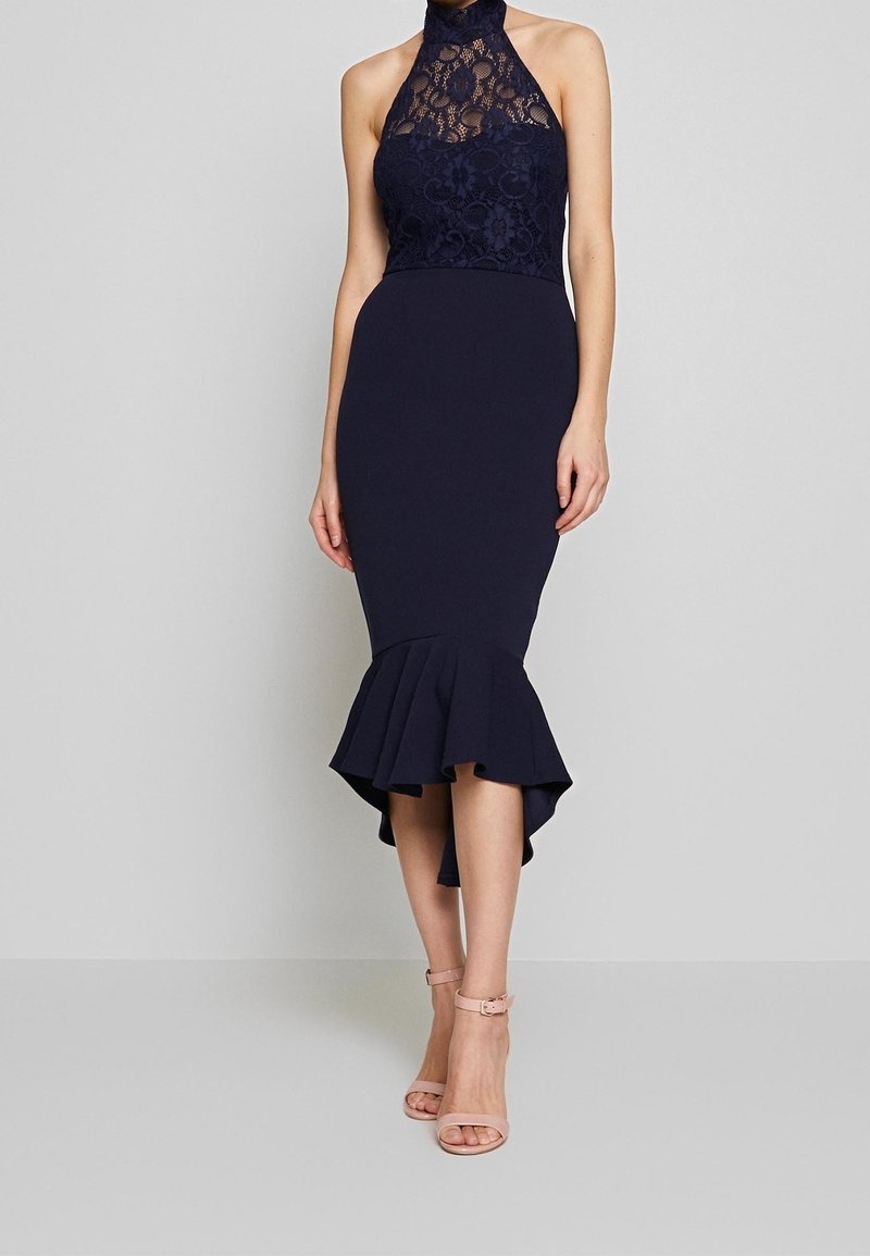 Femme portant une robe bustier en dentelle bleu marine avec un ourlet sirène et des talons nude à bride à la cheville, debout devant un fond uni.