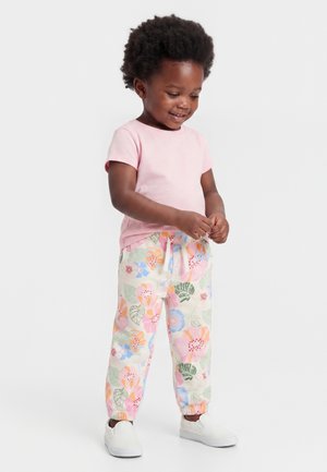 Toddler iført en pink t-shirt, blomstrede bukser med elastiske manchetter og hvide slip-on sko, stående mod en hvid baggrund.