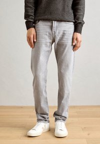 Blugi gri deschis cu o croială slim, având cinci buzunare și o ușoară decolorare. Combinați cu adidași albi pe un fundal din lemn.