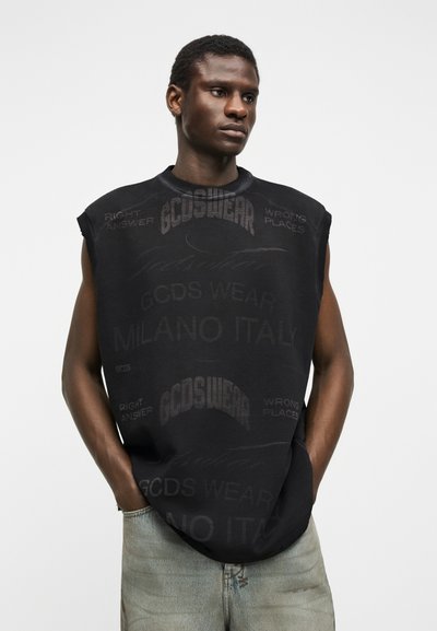 Νεαρός άνδρας που φορεί μαύρο αμάνικο μπλουζάκι με ξεθωριασμένο κείμενο "GCDS Wear Milano Italy" και ανοιχτό γκρι τζιν, στέκεται με τα χέρια στις τσέπες.