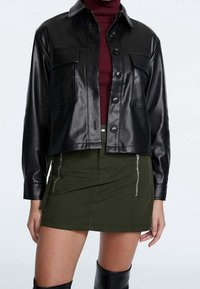 Veste en cuir noire coupée courte avec poches avant, col arrondi et fermetures à boutons, associée à une jupe vert olive à fermetures éclair latérales.