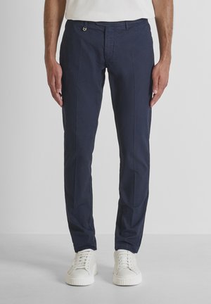 Homme portant un pantalon slim bleu marine et des baskets blanches, debout devant un fond gris clair uni.