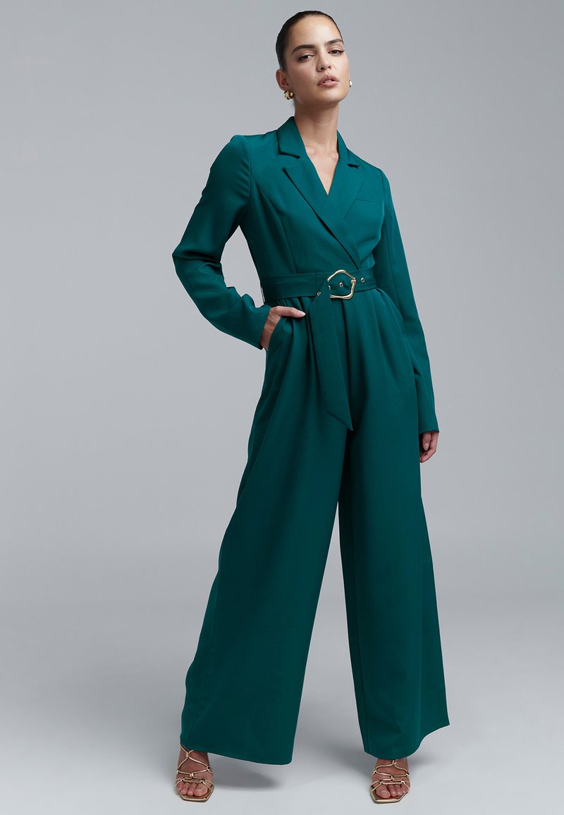 Teal jumpsuit with long sleeves, a tailored silhouette, a belted waist featuring a gold-tone buckle, and side pockets. Smooth fabric texture.