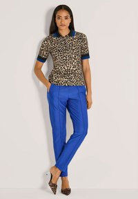 Vrouw met steil haar, gekleed in een poloshirt met luipaardprint, gedurfde blauwe tapered broek en hoge schoenen met luipaardprint.