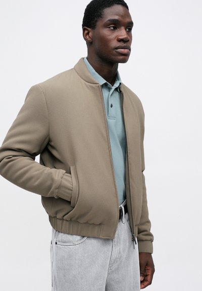 Ταπέ bomber μπουφάν με ριγωτούς καρπούς και γιακά, φορεμένο πάνω από ένα ανοιχτό μπλε polo, συνδυασμένο με ανοιχτό γκρι παντελόνι. Λείο υφή και φερμουάρ.