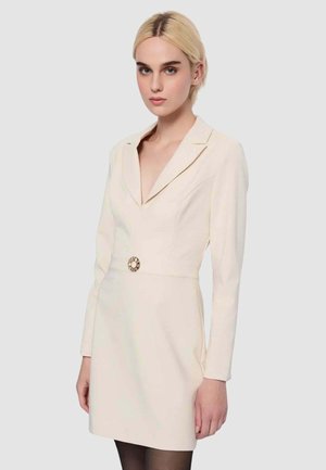 Donna bionda che indossa un abito blazer color crema con maniche lunghe, scollatura a V profonda e una fibbia circolare decorativa in vita.