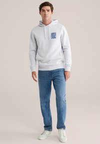 Felpa grigia chiara con grafica frontale, polsini a coste e tasca a marsupio, abbinata a jeans blu e sneakers chiare.