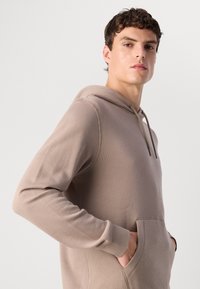 Jonge man met kort krullend haar, gekleed in een taupe hoodie, met zijn handen in de voorzak, die zelfverzekerd opzij kijkt tegen een effen achtergrond.