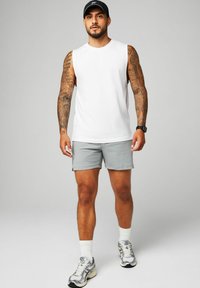 Weißes ärmelloses Shirt, hellgraue Shorts, Sportschuhe mit Akzenten und eine schwarze Mütze; Tattoos sichtbar auf den Armen. Einfaches Design, lässiger Stil.