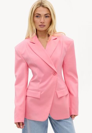 Mujer con una blazer rosa de gran tamaño con hombreras y vaqueros azul claro sobre un fondo blanco liso.
