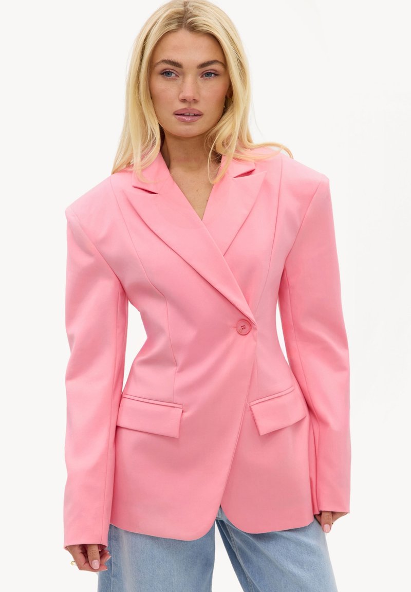 Femme portant un blazer rose oversize avec des épaules matelassées et un jean bleu clair sur un fond blanc uni.