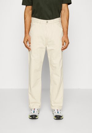 Mężczyzna nosi kremowe spodnie w prążki, połączone z ciemnozieloną koszulą i białymi trampkami z czarnymi i neonowo-żółtymi akcentami.