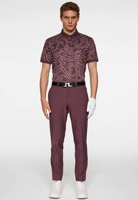 Męski strój golfowy składający się z bordowej koszulki polo z geometrycznym wzorem, bordowych spodni, białego paska i białych tenisówek. Biała rękawiczka na lewej ręce.