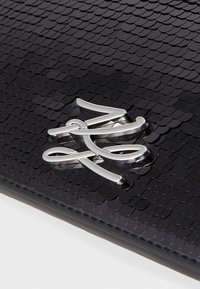Czarna torebka na ramię z teksturowanym wzorem w skale, zdobiona srebrnym metalowym akcentem w postaci logo. Gładkie krawędzie i elegancka forma.