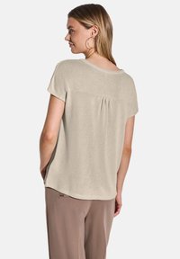 Svetle béžový krátký rukáv top s texturovaným povrchem, kulatým výstřihem a sklady na zadní straně. V kombinaci s hnědými kalhotami.