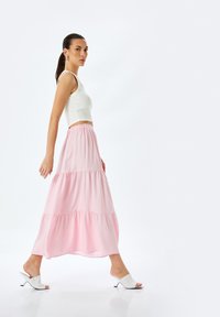 Gonna maxi a balze rosa con vita elastica, abbinata a un top corto bianco e sandali bianchi a ciabatta. Sfondo semplice.