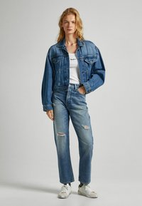 Jeans jakna v srednji modri barvi z oversized krojem, prsni žepi in vidnim šivanjem. V kombinaciji s svetlo modrimi uničenimi kavbojkami in belimi supergami.