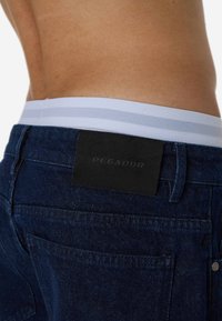 Τζιν σε σκούρο μπλε χρώμα με υφασμάτινη μέση που έχει λευκές ρίγες, και μία μαύρη δερμάτινη ετικέτα με ανάγλυφο το "PEGADOR" στο πίσω μέρος.