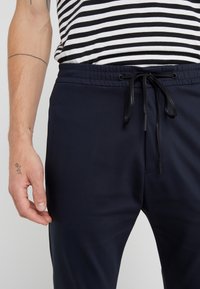 Man in een marineblauwe broek met zwart trekkoord en een gestreept zwart-wit shirt, zichtbare tatoeages op de linker onderarm en hand.