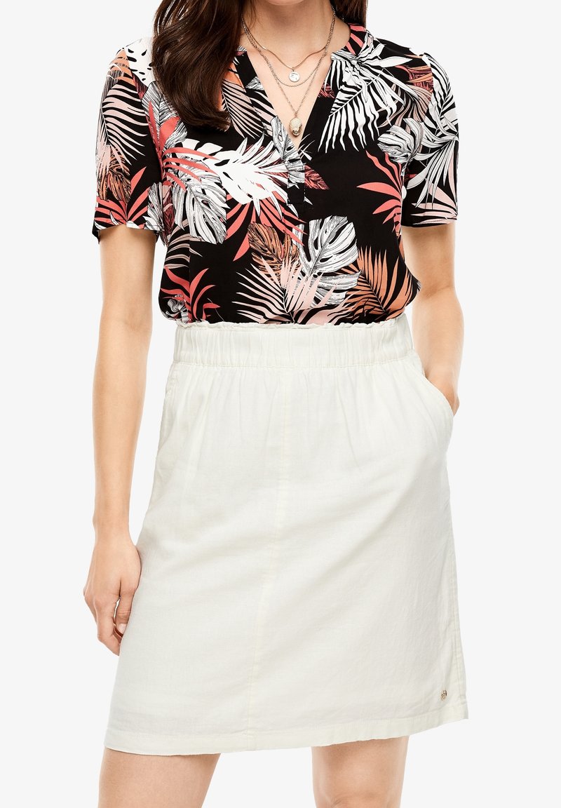 Femme portant un chemisier noir à manches courtes à motif feuilles corail, rentré dans une jupe blanche taille haute, longueur genoux, avec poches.