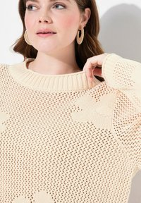 Maglione di colore crema lavorato a maglia con una trama larga e scollatura a coste. Presenta dettagli testurizzati e una vestibilità comoda. È visibile un paio di orecchini a cerchio.