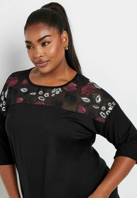 Musta paita, jossa on 3/4-hihat, läpinäkyvä verkkoyläosa, joka on koristeltu brodeeratuilla ruusuilla ja lehdillä burgundin, mustan ja hopean sävyissä.