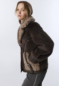 Jeune femme portant une veste en fourrure synthétique brun foncé avec un grand col et une boucle sur le côté, posant de profil contre un fond uni.