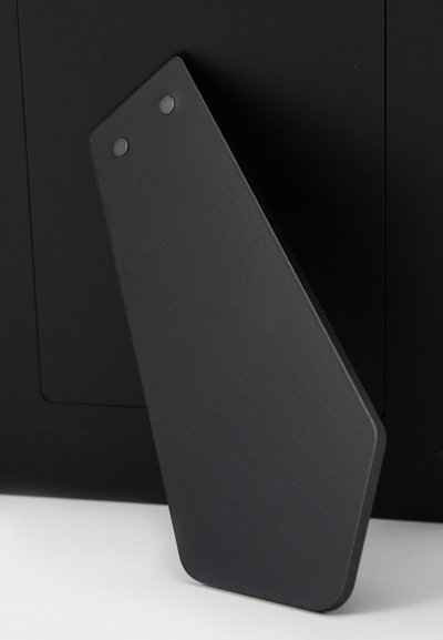 Czarna matowa etykieta o geometrycznym, kanciastym kształcie i zaokrąglonych rogach. Posiada dwa okrągłe otwory blisko górnej krawędzi do mocowania.