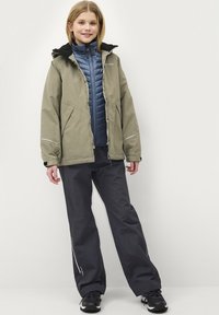 Olivengroene buitenjas met zwarte capuchon, blauwe geïsoleerde binnenvest, grijze broek en zwarte schoenen. Bevat reflecterende accenten en ritsen.