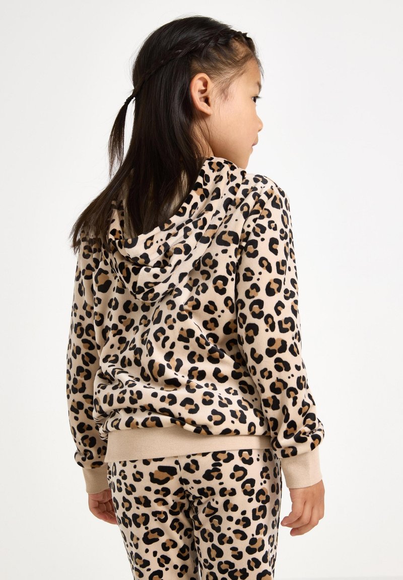 Fille aux cheveux foncés portant un hoodie à imprimé léopard beige et un pantalon assorti, debout et légèrement tournée vers la droite devant un fond uni.