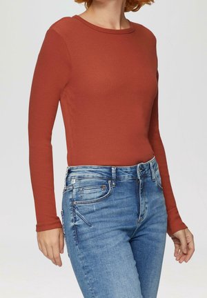 Personne portant un haut ajusté à manches longues couleur rouille et un jean bleu en denim avec poches avant et arrière visibles.