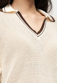 Pull-over en tricot avec un col en V et un col contrastant marron et blanc. Couleur beige clair avec un motif texturé en maille ouverte.
