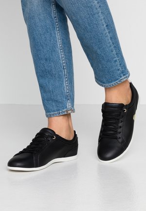 Personne portant des baskets noires à lacets avec des semelles blanches et un jean bleu, debout sur un sol blanc sur fond uni.