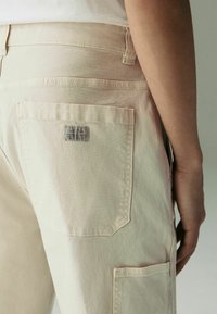 Beige cargo pants featuring multiple pockets, a textured fabric, and a stitched label on the back pocket, highlighting utilitarian design.