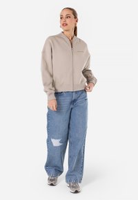 Beige zip-up hettegenser med ribbede mansjetter, kombinert med lyseblå vide bukser med en slitt lapp. Modellen har på seg grå joggesko.