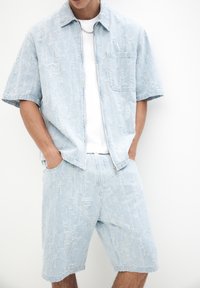 Vaaleansininen denim-paitapusero lyhyillä hihoilla ja taskulla, yhdistettynä vastaaviin shortseihin. Molemmat vaatteet ovat teksturoitua kangasta ja niissä on repaleiset helmat.