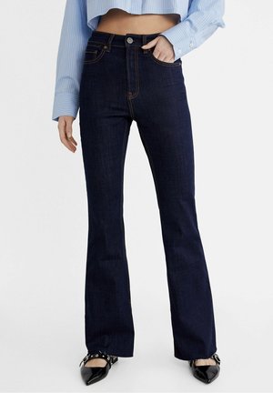 Kobieta w ciemnoniebieskich jeansach o wysokim stanie i kroju bootcut, jasno-niebieskiej krótkiej koszuli oraz czarnych, szpiczastych butach z paskami wokół kostek.