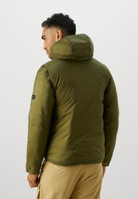 Veste doudoune vert olive avec un col montant, présentant une texture lisse et une étiquette noire sur la manche. Conception à capuche avec des poignets élastiques.