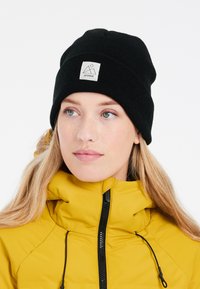 Berretto lavorato a maglia nero in tessuto a coste, dotato di un'etichetta frontale con un design geometrico. Indossato con un giubbotto giallo imbottito.