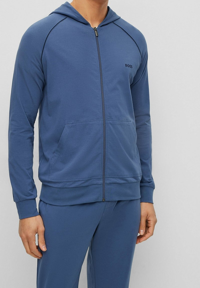 Homme portant un sweat à capuche bleu zippé et un pantalon assorti, avec des poches avant et le logo "BOSS" sur la poitrine.