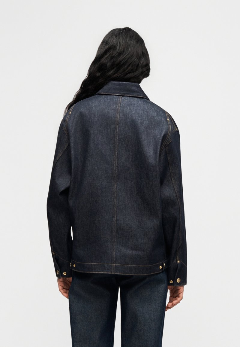 Veste en denim foncé avec une coupe ajustée, un grand revers et des boutons en laiton. La vue de dos montre des coutures contrastées et une silhouette décontractée.